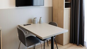 A modern, minimalistic room with a flat-screen TV mounted on the wall, a wooden cabinet, a dining table with two chairs, and a remote control on the table.
