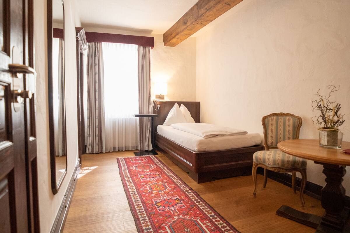 A hotel room with a wooden floor, a large bed, a chair, a small table, and a red patterned rug.