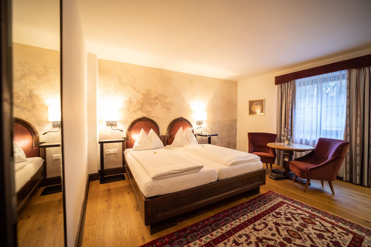 A hotel room with a double bed, two bedside tables with lamps, a small round table with a chair, and a large window with curtains.