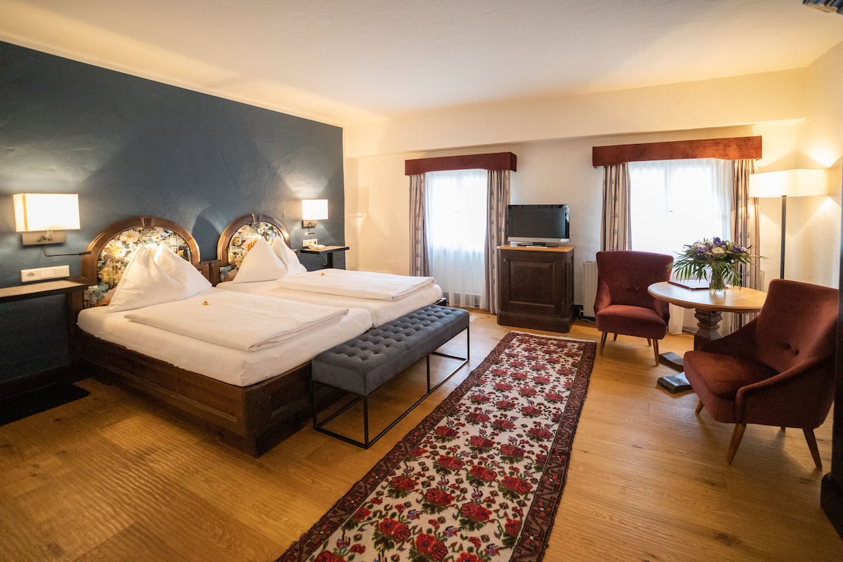 A hotel room with a double bed, a bench, a TV, a desk, a chair, and a small table with a vase of flowers.