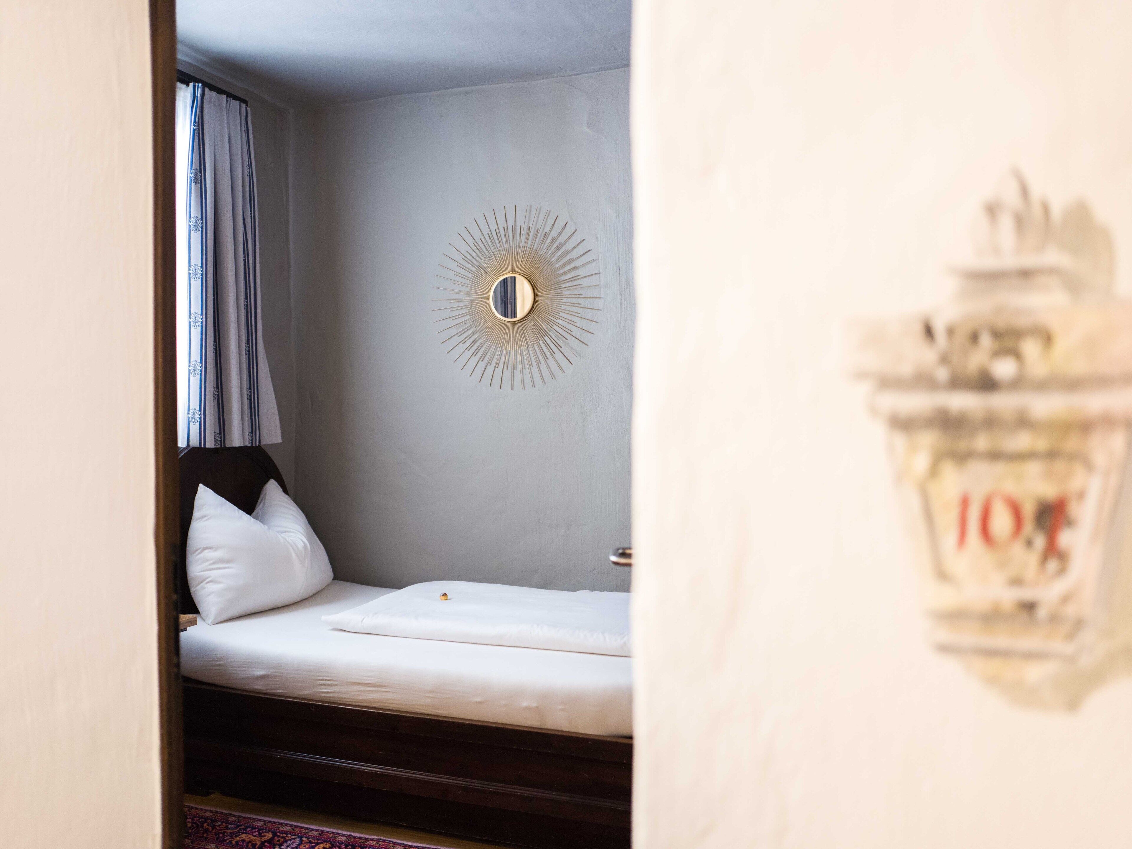 Ein kleines, ordentlich bezogenes Bett in einem Zimmer mit einem Wandspiegel und einem Vorhangfenster.