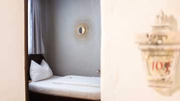 Ein kleines, ordentlich bezogenes Bett in einem Zimmer mit einem Wandspiegel und einem Vorhangfenster.