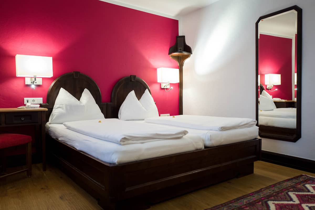 A double bed with white linens, a wooden headboard, two bedside lamps, a red wall, a mirror, a desk with a chair, and a patterned rug.
