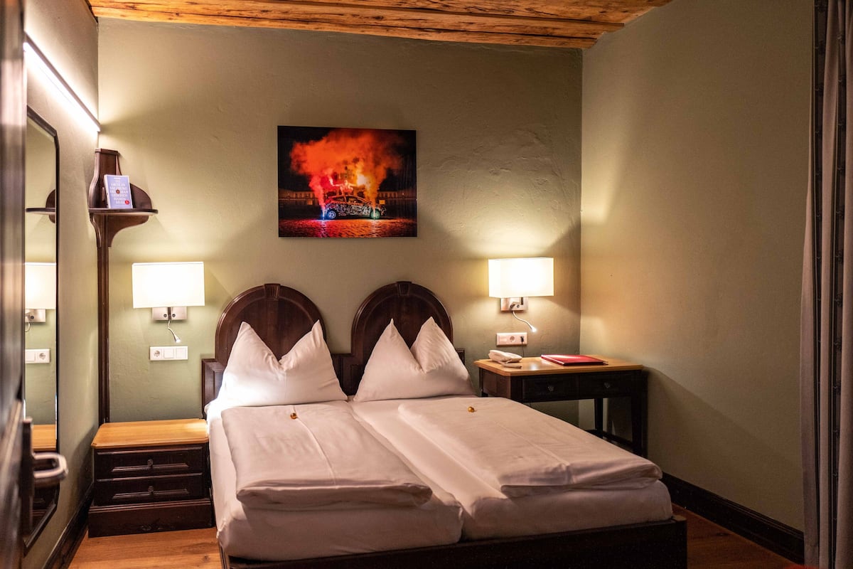 A hotel room with a bed, bedside tables, lamps, a mirror, and a painting of a fire on the wall.