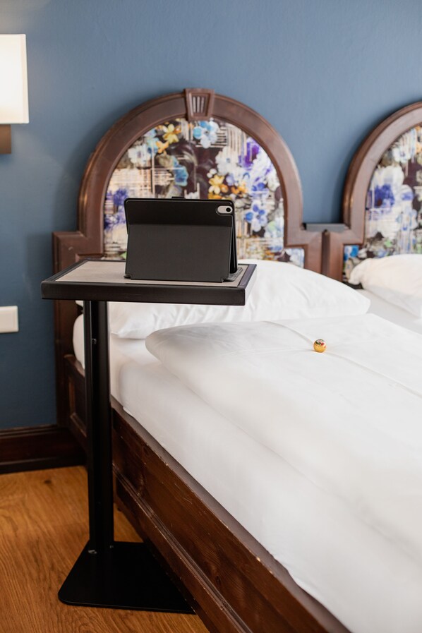 A laptop on a bedside table in a hotel room with a wooden bed frame and floral wall decor.