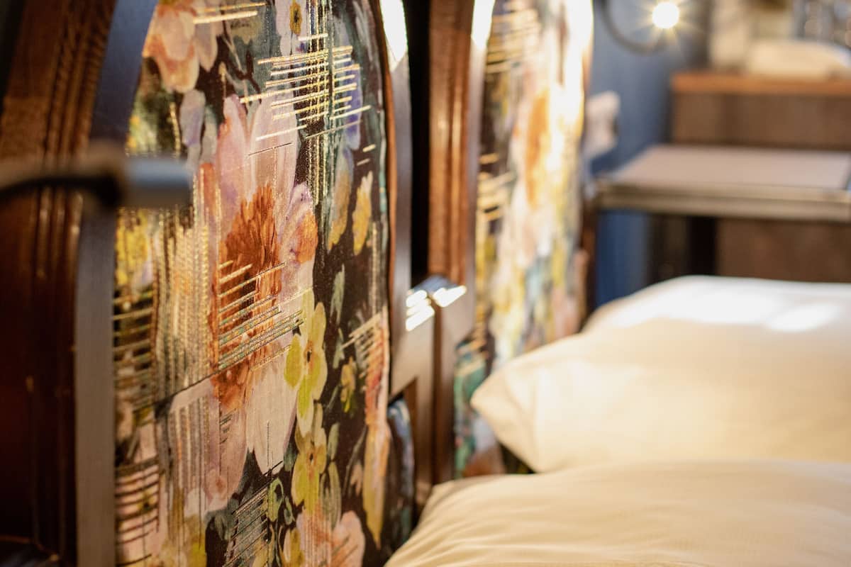 A hotel room with a bed, floral-patterned headboard, a bedside table, and a lamp.