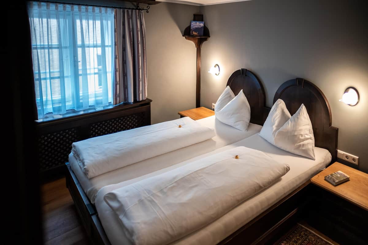 A double bed with white linens, a bedside table with a book, a nightstand with a lamp, and a window with curtains.
