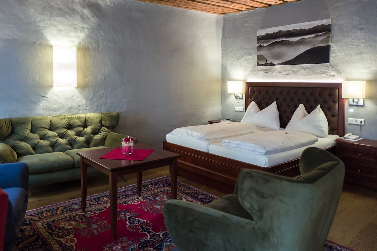 A hotel room with a large bed, a green sofa, a small table with a red placemat, and a patterned rug.