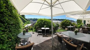 An outdoor patio with tables and chairs under large umbrellas, surrounded by lush greenery and a view of buildings in the distance. - Altstadthotel Stadtkrug (Salzburg)
