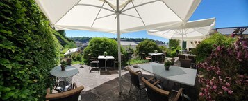 Un patio al aire libre con mesas y sillas bajo grandes sombrillas, rodeado de abundante follaje y vistas a edificios en la distancia.