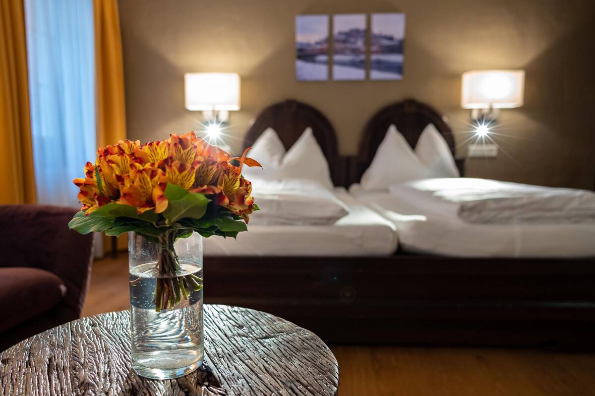 A hotel room with a bed, two bedside lamps, a vase with flowers on a round table, and a view of a snowy landscape through the window.