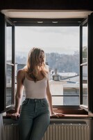 Una persona de pie junto a una ventana con vistas a edificios y árboles.