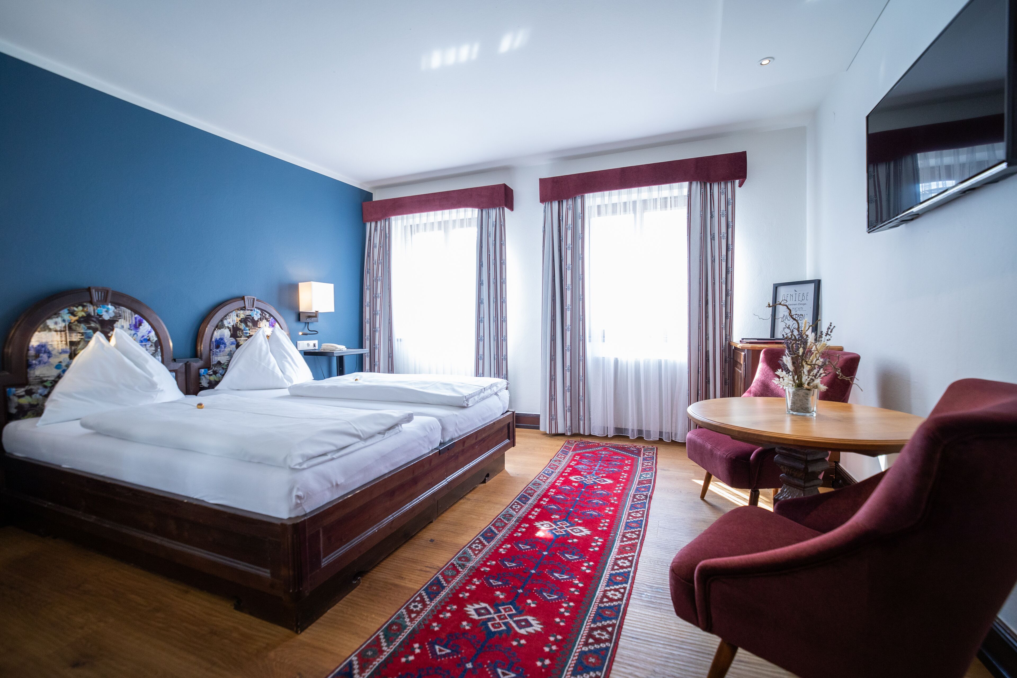 A hotel room with a large bed, a round wooden table, a red armchair, and a TV mounted on the wall.