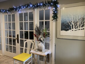 A door with frosted glass panels, a white chair with a yellow cushion, a small table with a vase of dried plants, and a framed picture on the wall. - Hotel & Suites Les Laurentides Saint Sauveur (Saint-Sauveur)