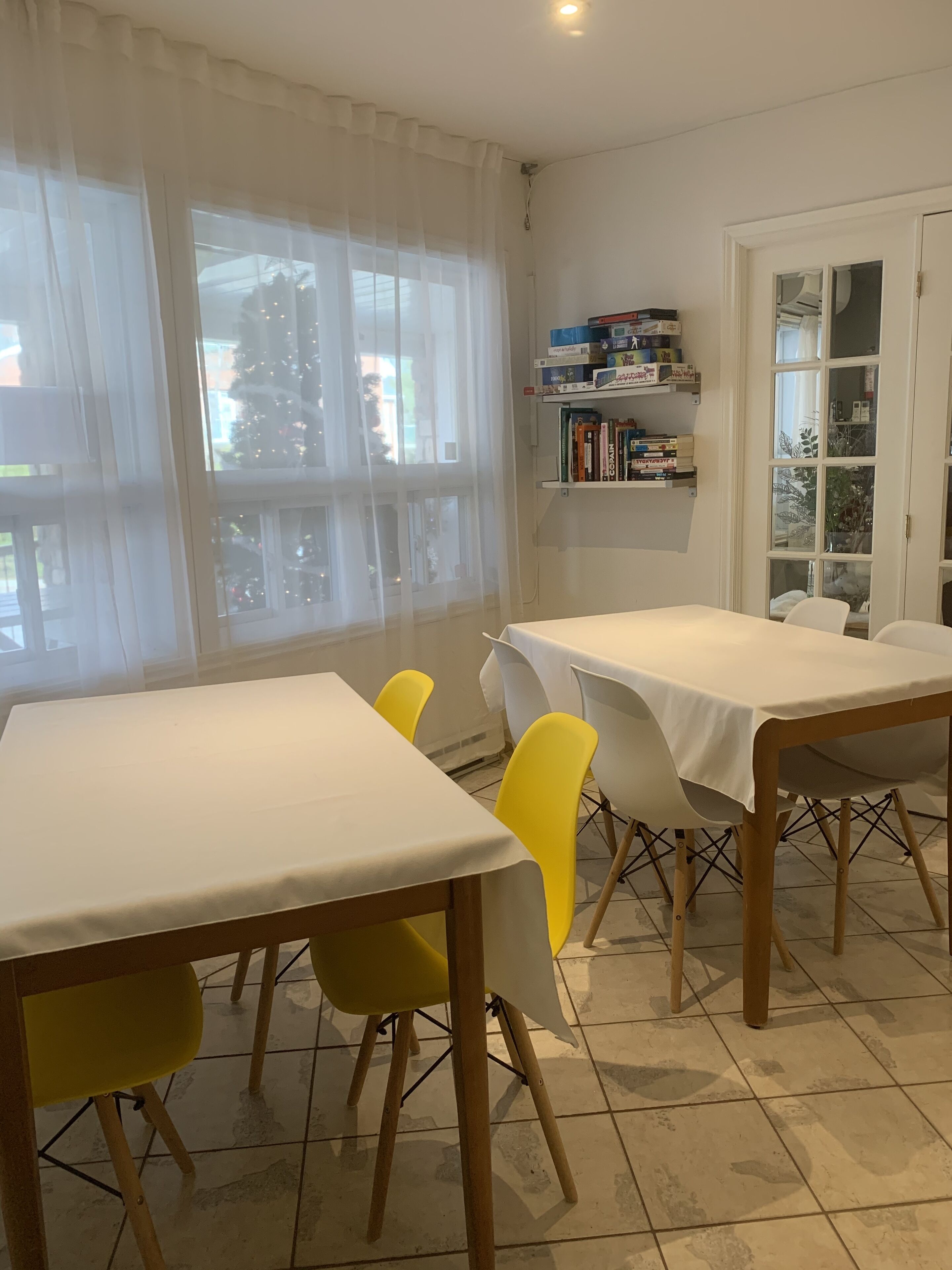 Un comedor con mesa blanca y sillas amarillas, una estantería con libros, y una ventana con cortinas traslúcidas.