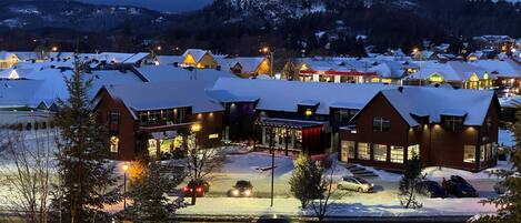 A snow-covered resort with multiple buildings, a central open area with seating, and a view of snow-covered mountains in the background.