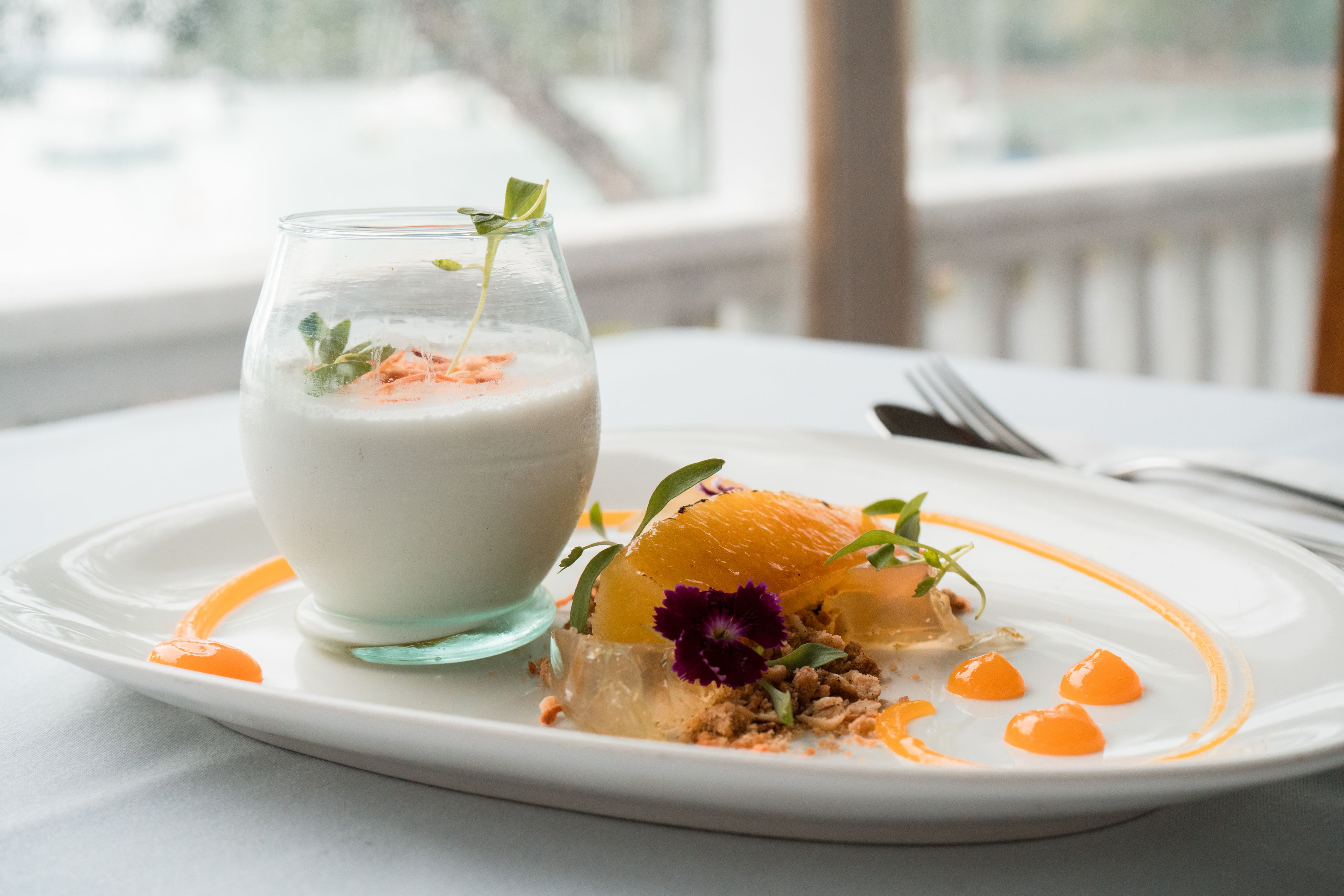 A dessert plate with a glass of a creamy beverage, garnished with orange slices and edible flowers.