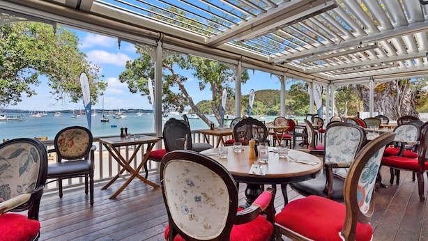 Un área de comedor al aire libre con mesas y sillas, con vista a una bahía con barcos.