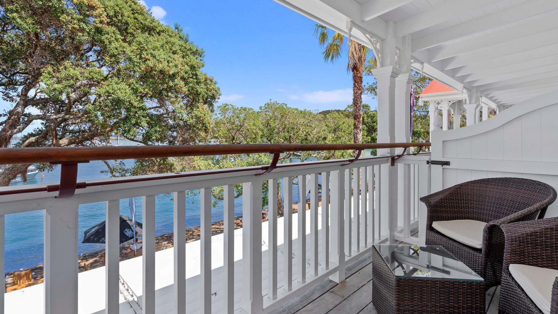 Ein überdachter Balkon mit Korbmöbeln, weißen Geländern und Blick auf Meer und Bäume.