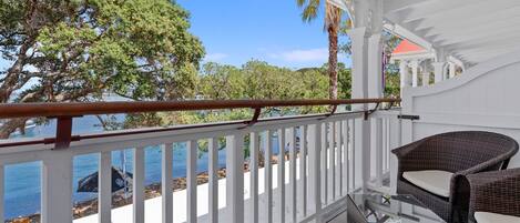 Ein überdachter Balkon mit Korbmöbeln, weißen Geländern und Blick auf Meer und Bäume.