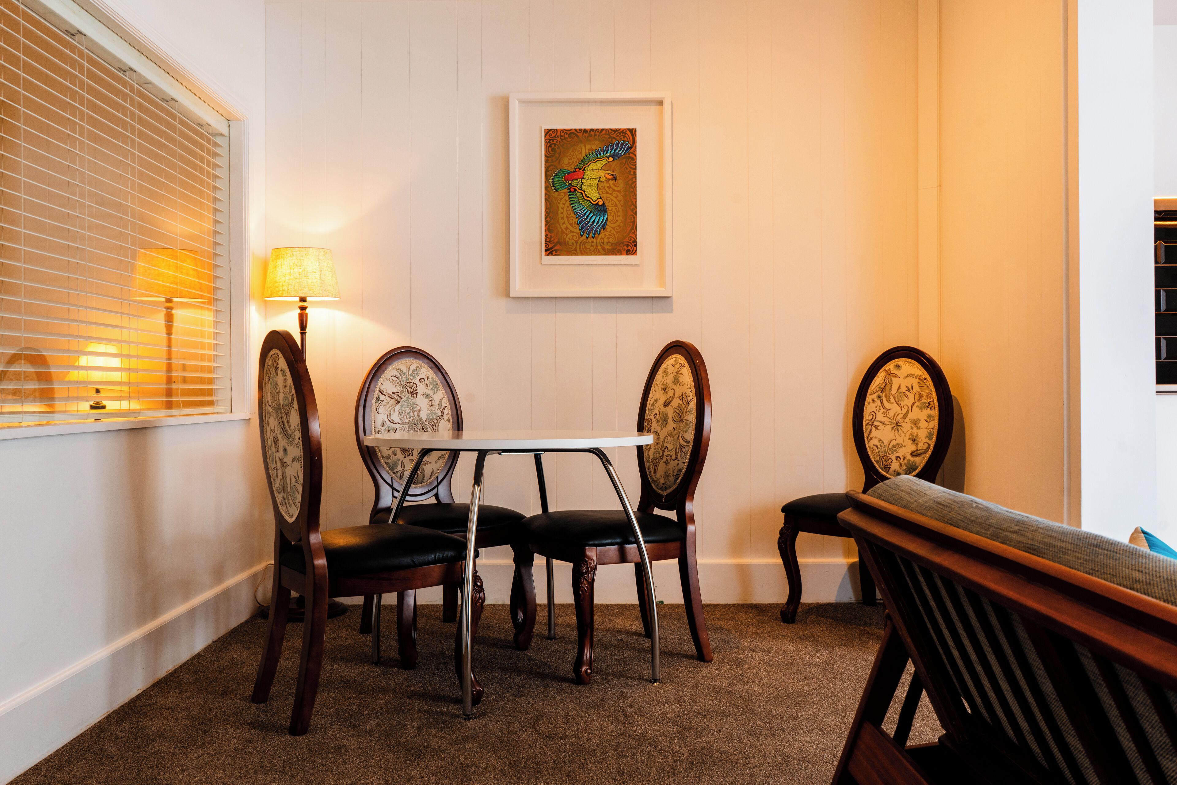 Un comedor con una mesa y cuatro sillas, una obra de arte enmarcada en la pared y una ventana con persianas.