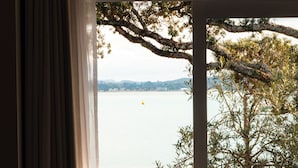 Vista a través de una ventana con cortina, mostrando una masa de agua, un árbol y una cadena montañosa distante.