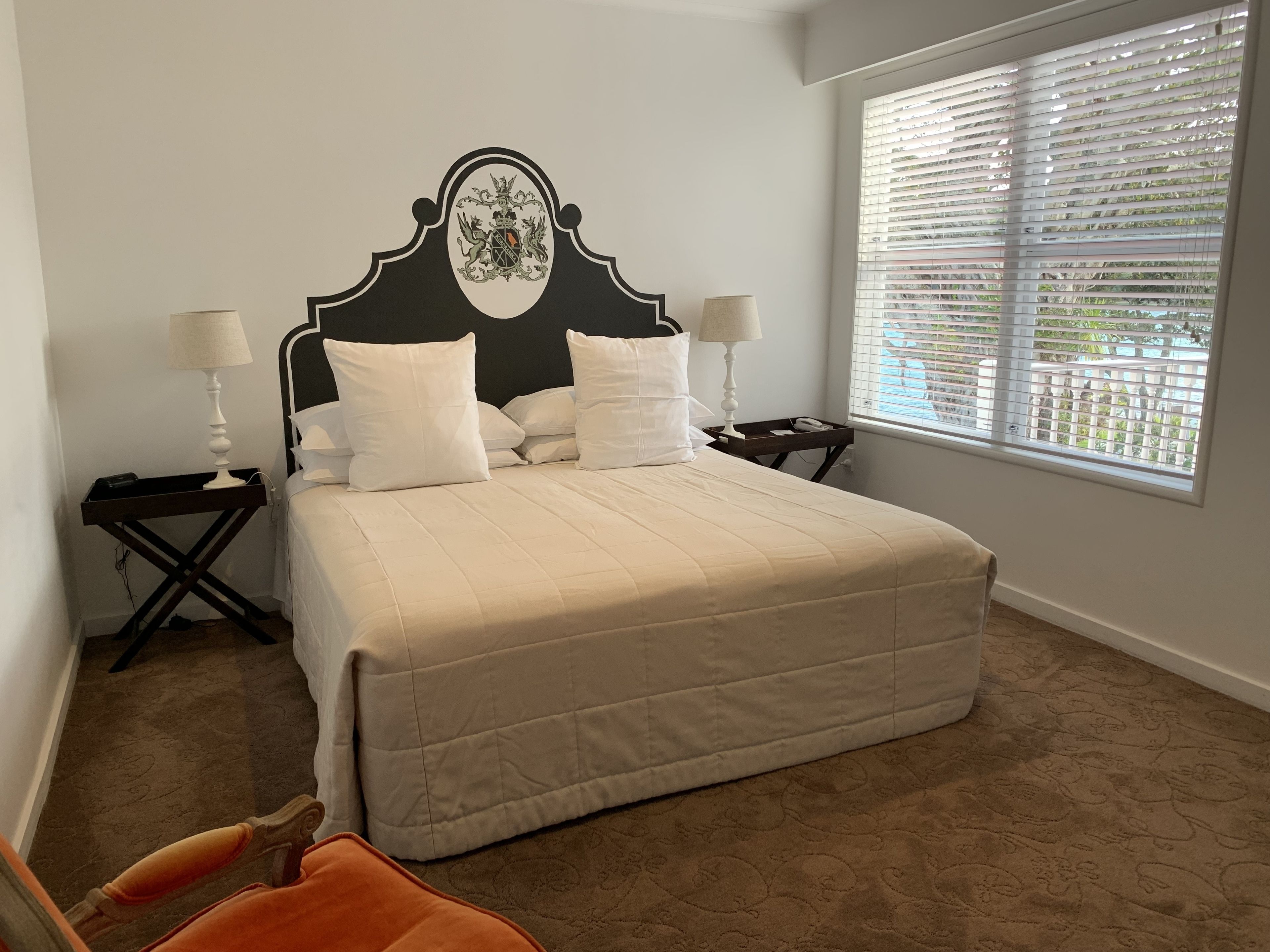 A neatly made bed with a headboard, two bedside tables with lamps, a window with blinds, and a patterned carpet.