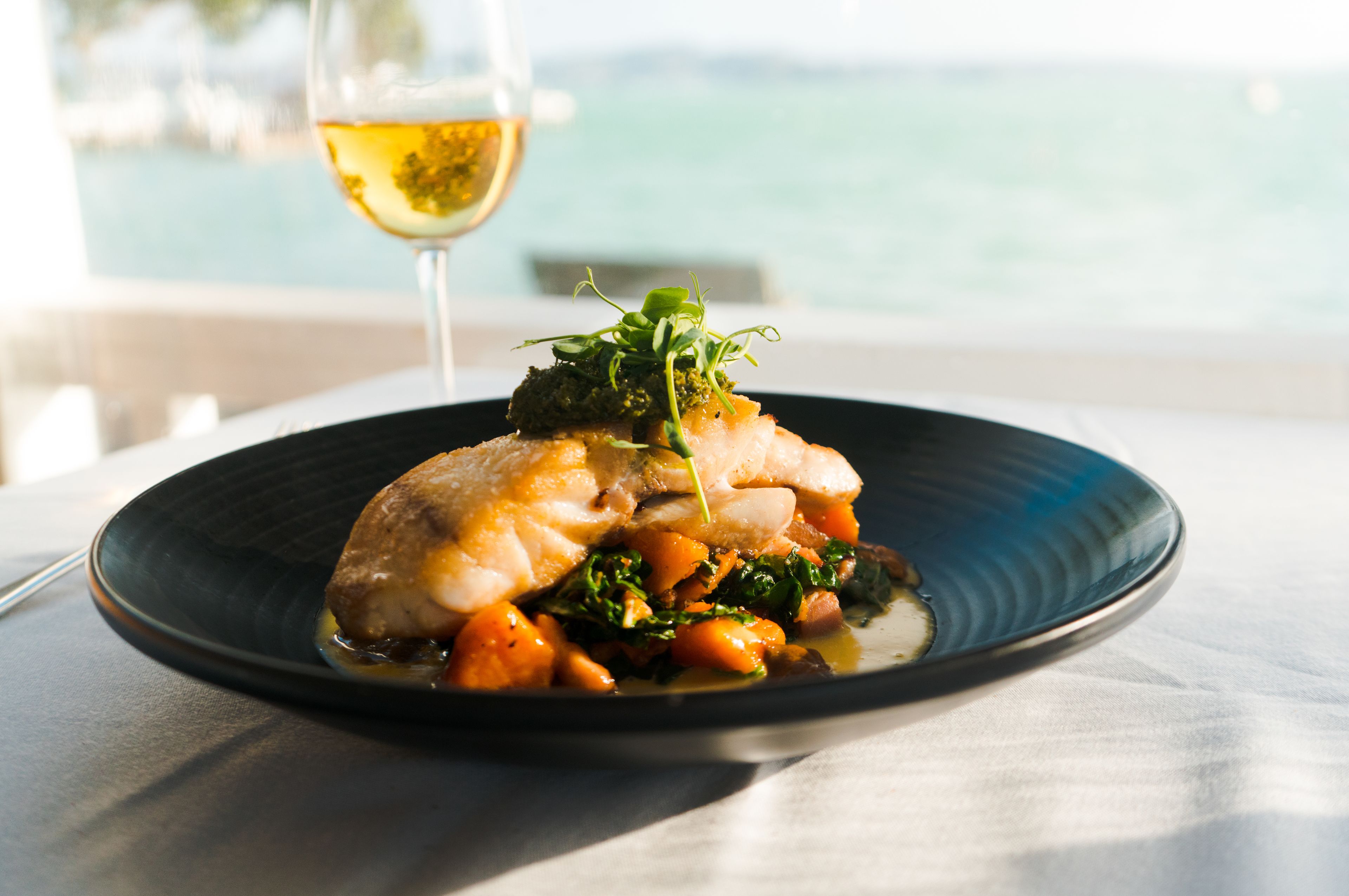 A plate of food with a glass of wine on a table by a window overlooking a body of water.