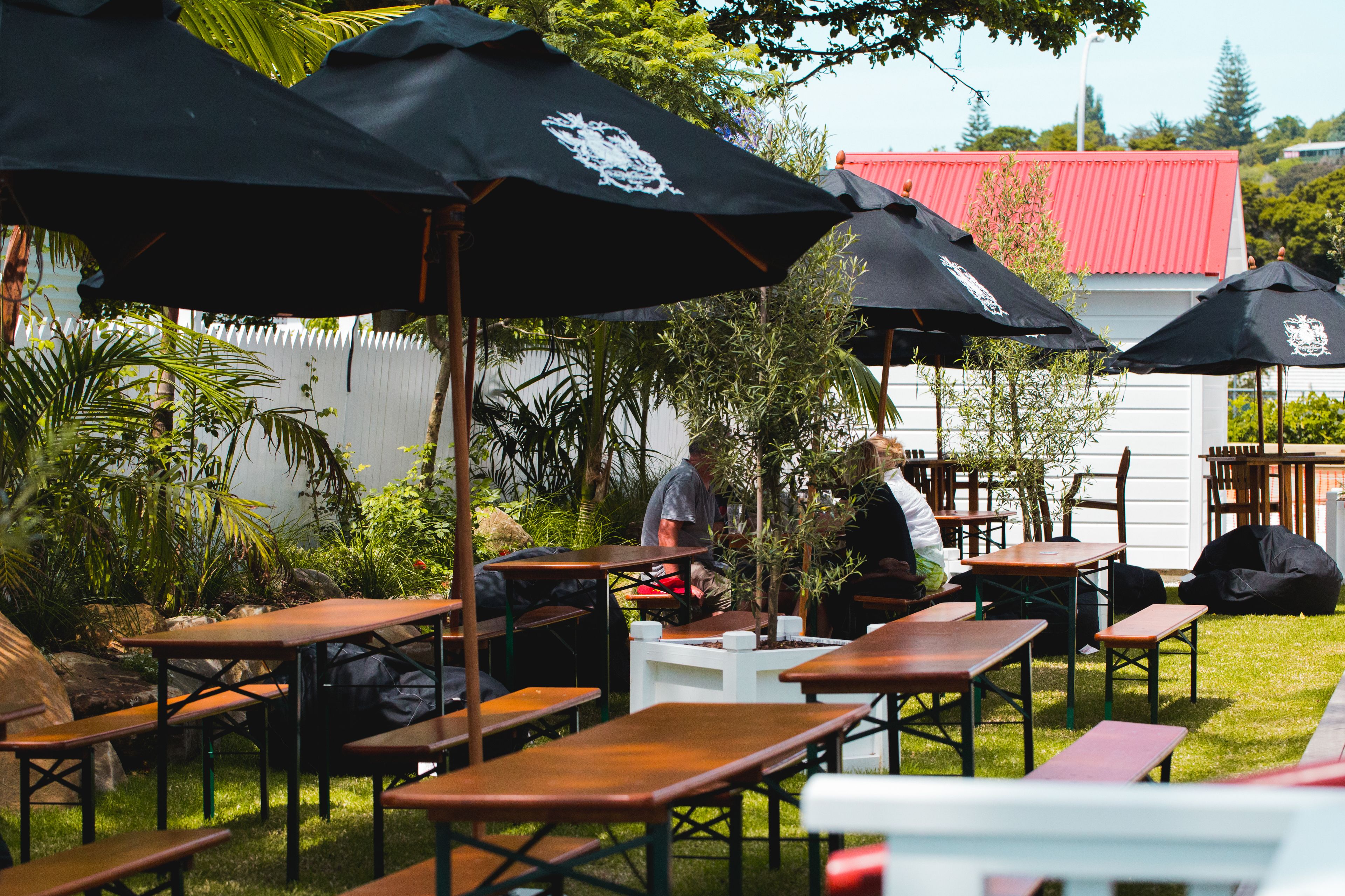 An outdoor dining area with wooden tables and black umbrellas, surrounded by greenery and a white fence.