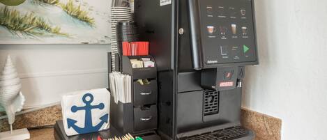 Una máquina de café Nescafé con pantalla digital, una pila de vasos desechables y una bandeja de servilletas de colores.