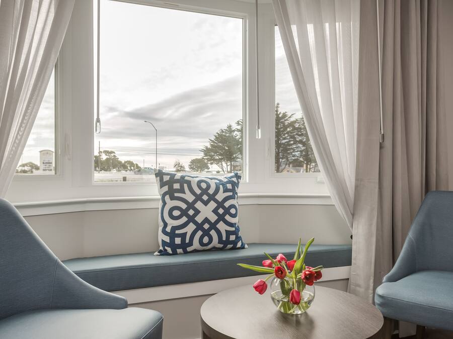 A room with a view of the city, a blue sofa, a small round table with a vase of red tulips, and a patterned pillow on the window seat.