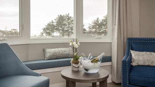 A room with a blue sofa, a small table with a plant and a champagne bucket, and a view of trees through large windows.
