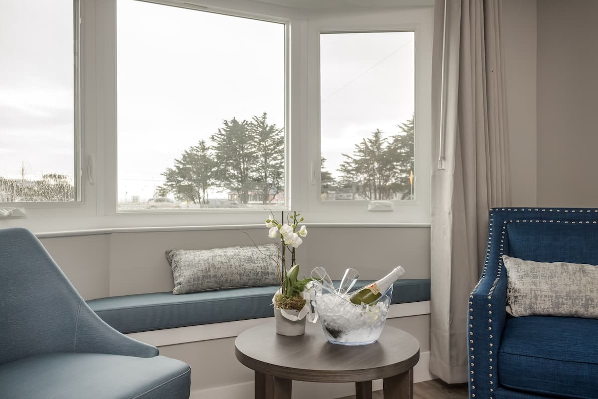 A room with a blue sofa, a small table with a plant and a champagne bucket, and a view of trees through large windows.