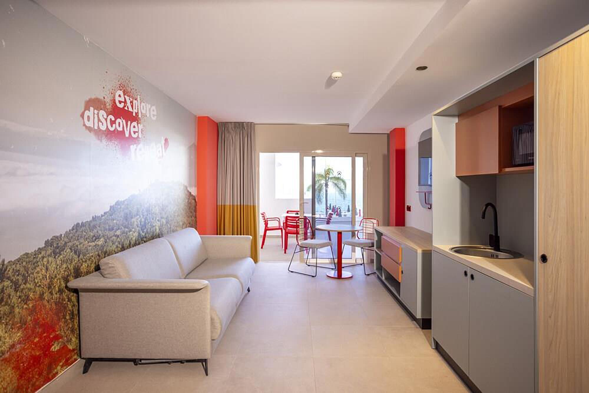 Une chambre d’hôtel moderne dotée d’un canapé, d’un coin repas avec des chaises, d’un coin cuisine et d’une fresque murale.