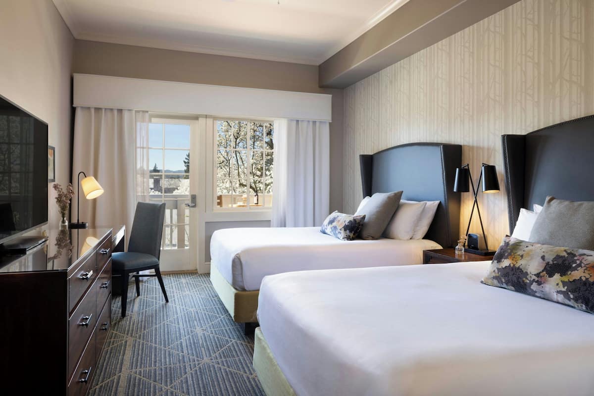 A hotel room with two beds, a desk with a TV, a chair, and a view of trees through the window.