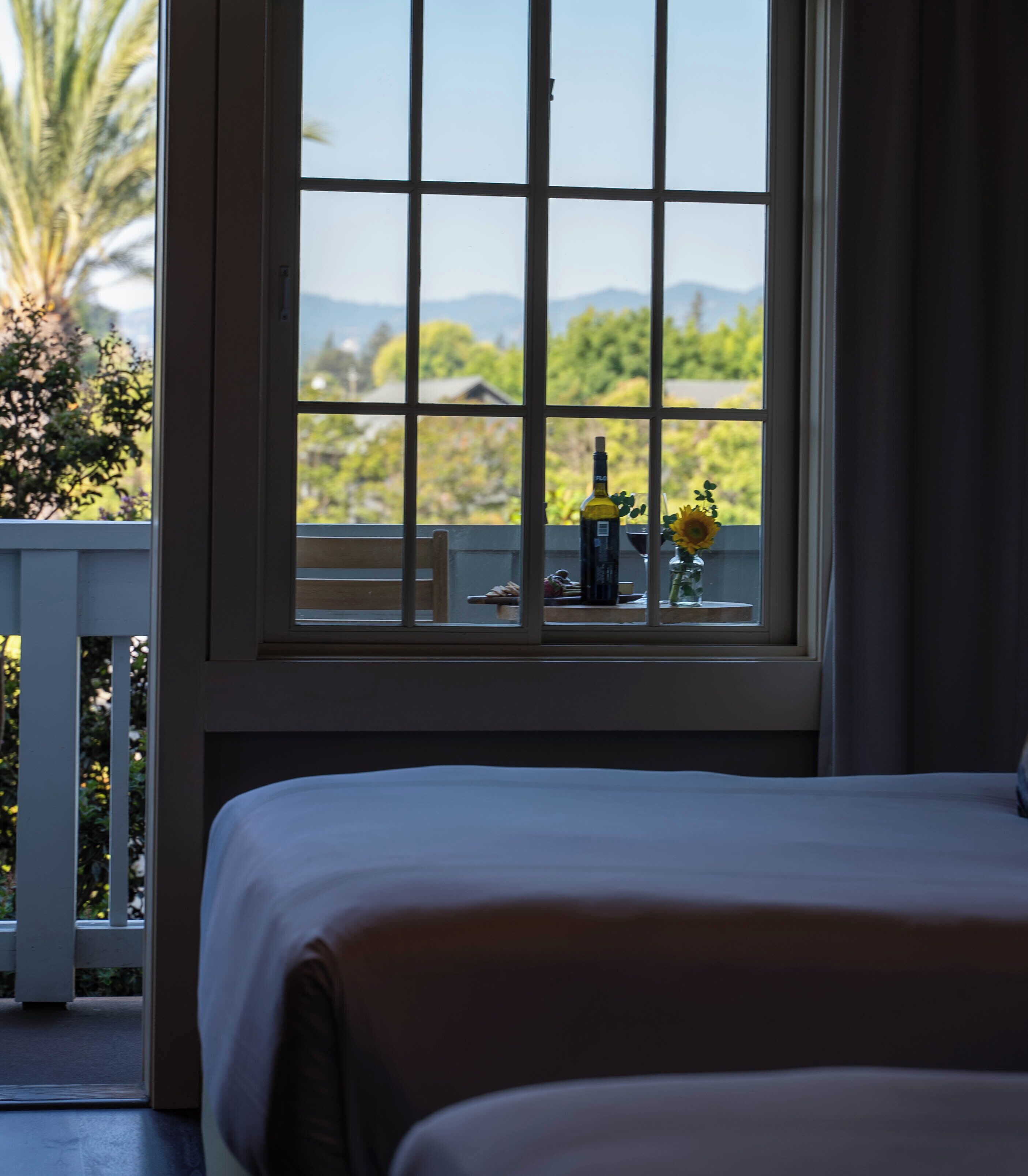 A hotel room with a bed, a window with a view of greenery and mountains, a bottle of wine, and a vase with flowers.