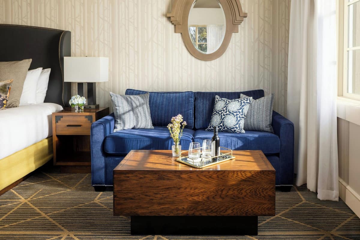 A hotel room with a bed, a blue sofa, a wooden coffee table, a nightstand, a lamp, and a mirror.