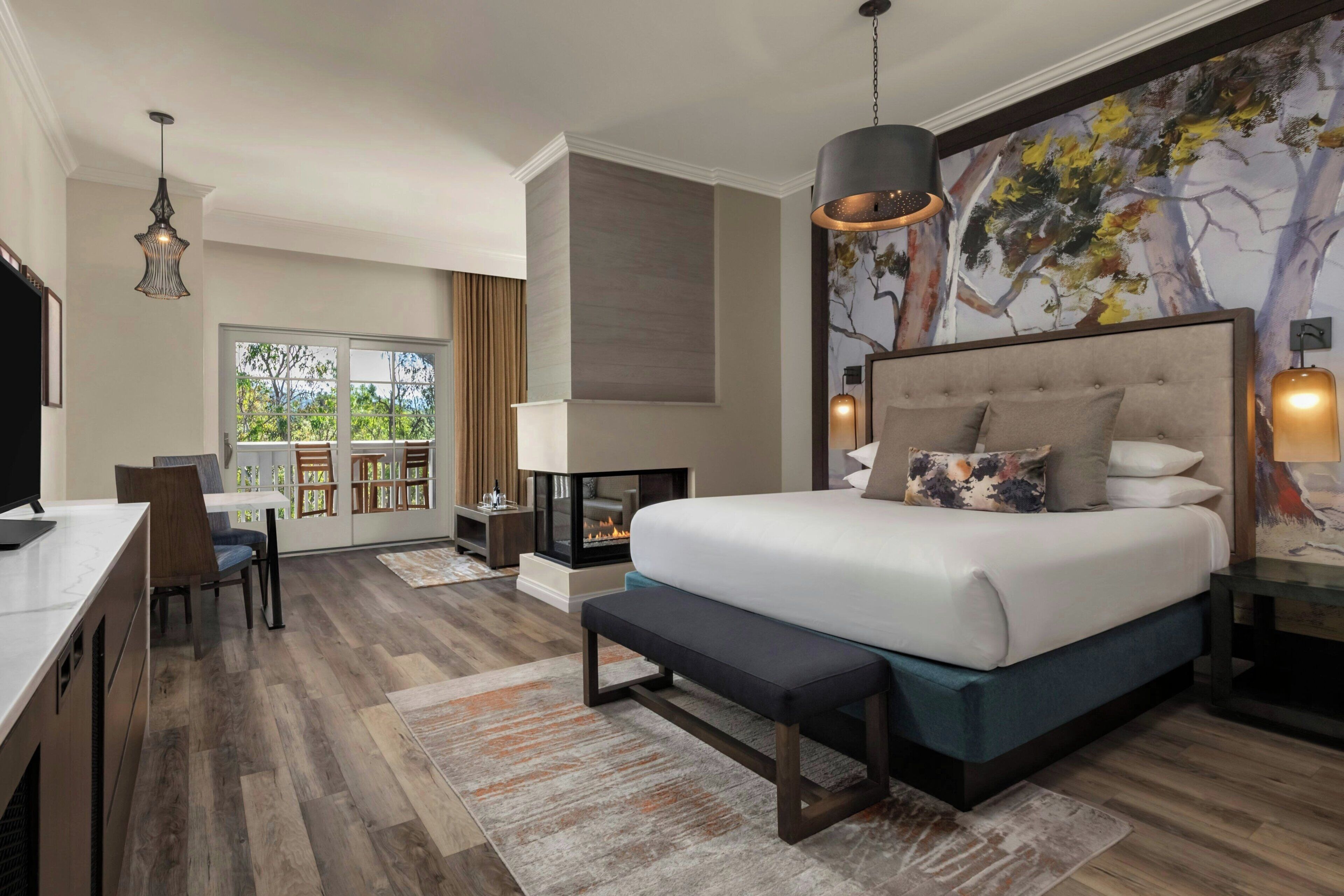A modern hotel room with a large bed, a desk, a fireplace, and a view of trees.