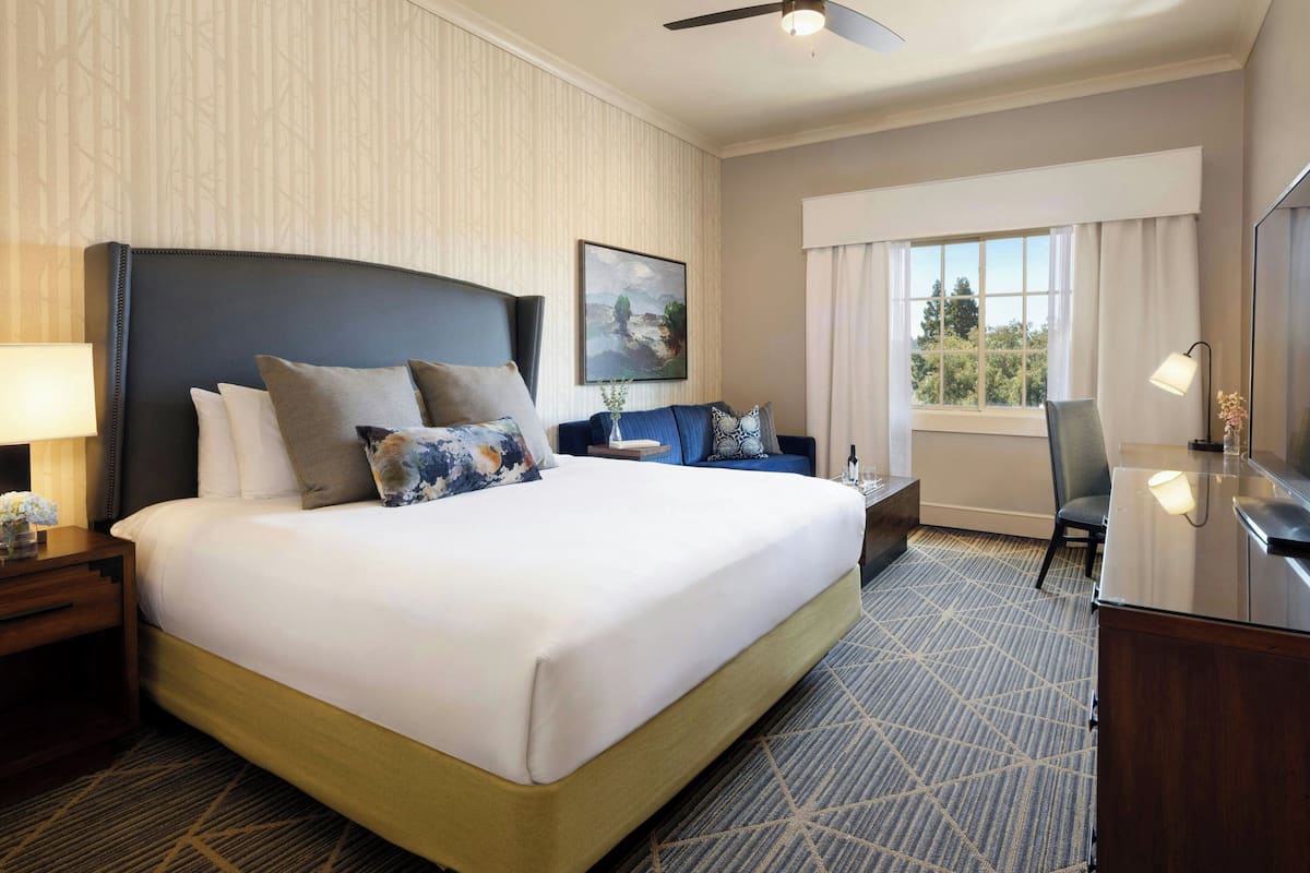 A hotel room with a large bed, a desk, a chair, a window with curtains, and a ceiling fan.