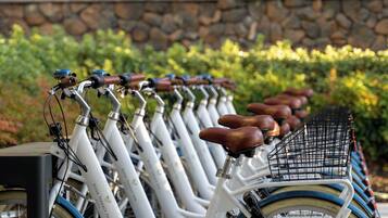 A row of white bicycles with brown seats and baskets, parked against a stone wall with greenery in the background.