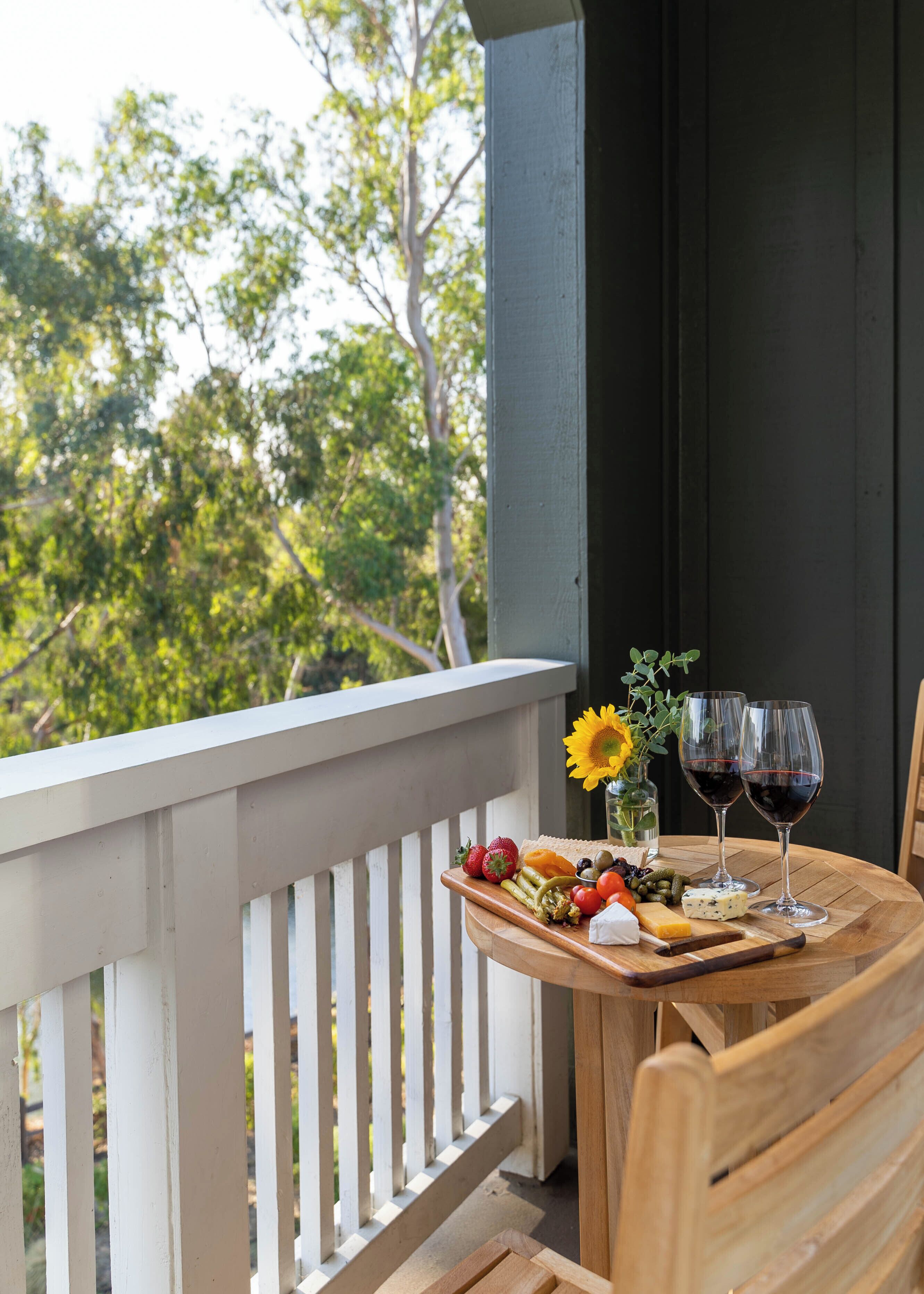 A wooden table with a fruit platter, two wine glasses, and a sunflower on a balcony with a white railing and a dark wall.