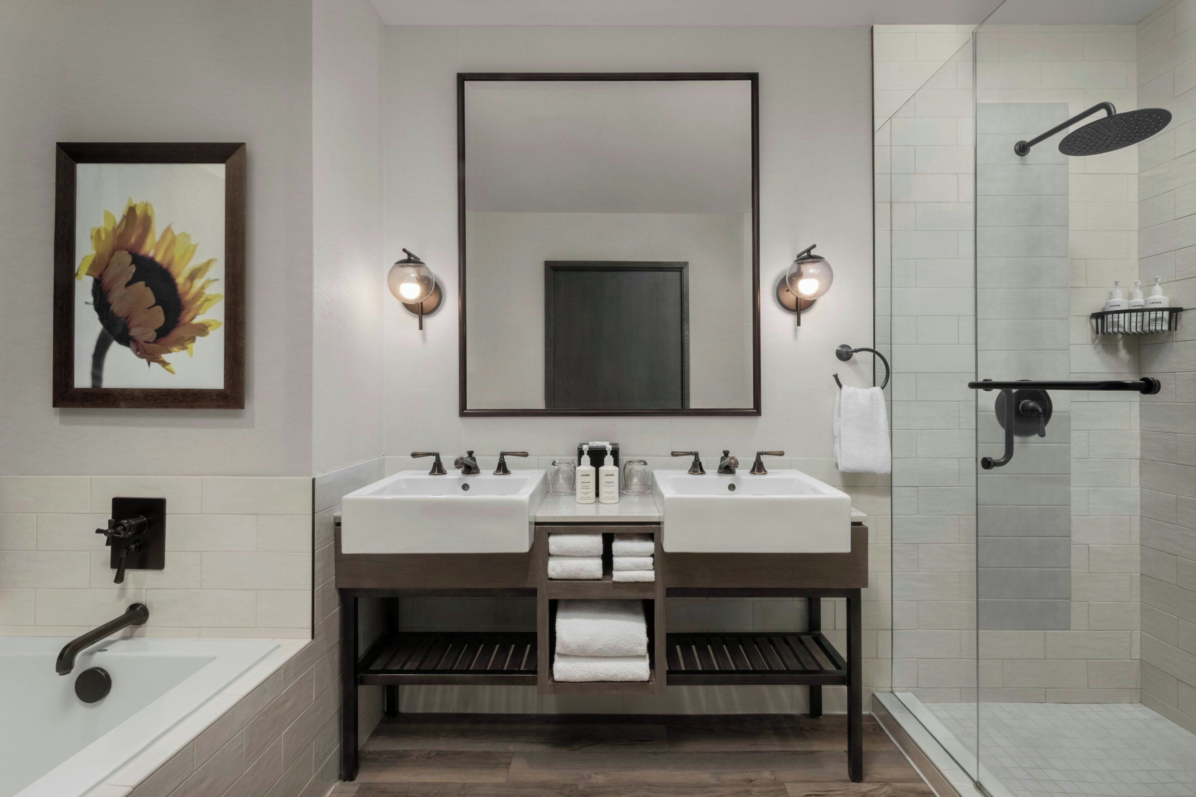 Une salle de bain moderne avec deux lavabos, un grand miroir, des appliques murales, une douche et une œuvre d’art encadrée.