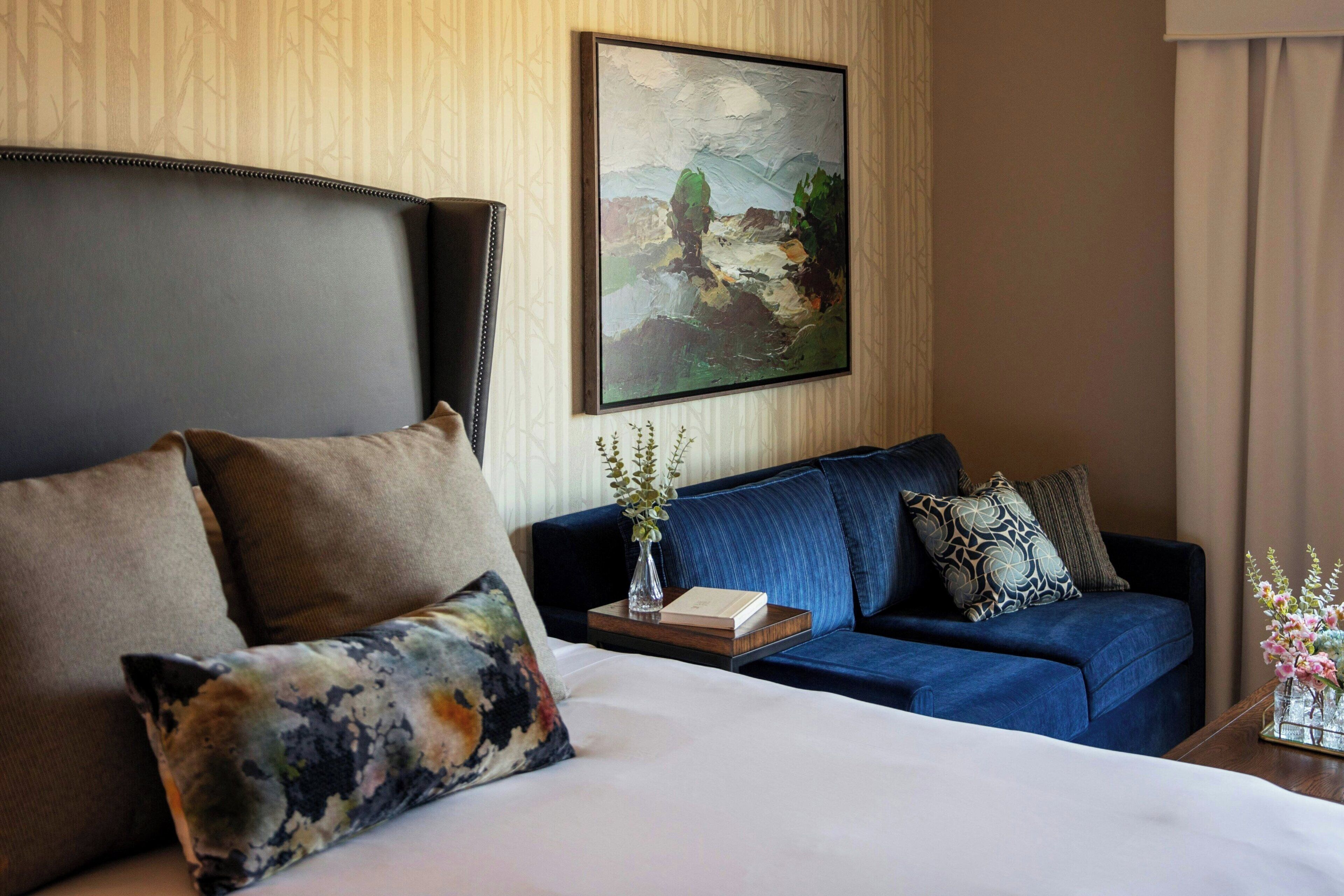 A hotel room with a bed, a sofa, a painting on the wall, and a vase with flowers on a side table.