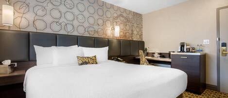 A hotel room with a large bed, a headboard, a bedside table with a lamp, a small desk with a chair, a minibar, and a wardrobe.