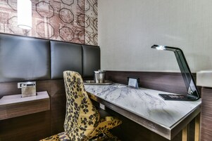 Una habitación de hotel moderna con un escritorio de mármol, un sillón cómodo, una mesita de noche con lámpara y un papel tapiz estampado.