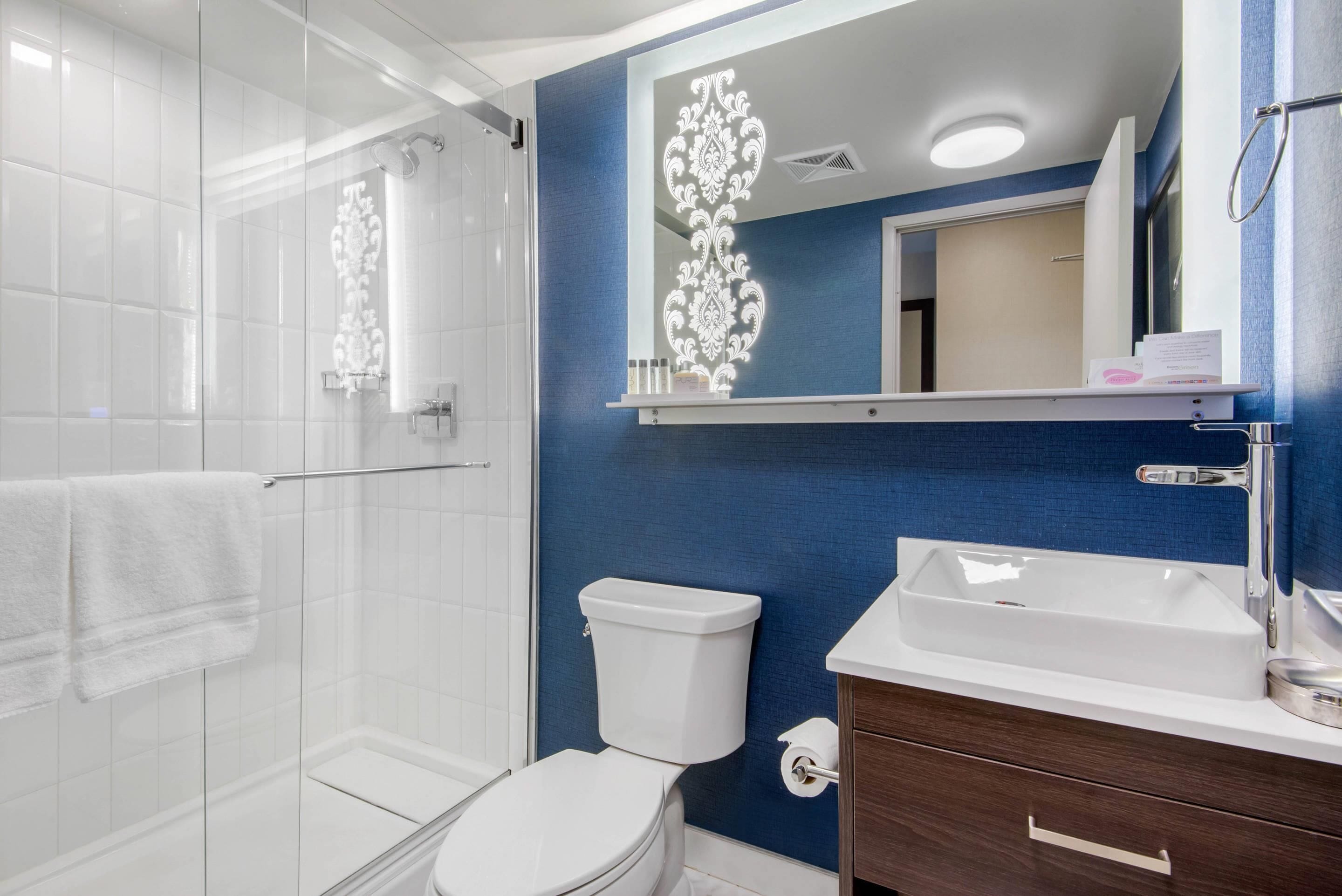 A modern bathroom with a glass shower enclosure, a white toilet, a blue accent wall, a white sink, and a vanity with a mirror.