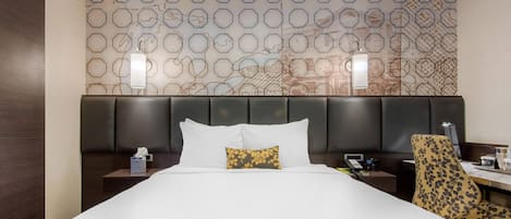 A hotel room with a large bed, a headboard with a geometric pattern, a nightstand with a lamp, and a desk with a chair.
