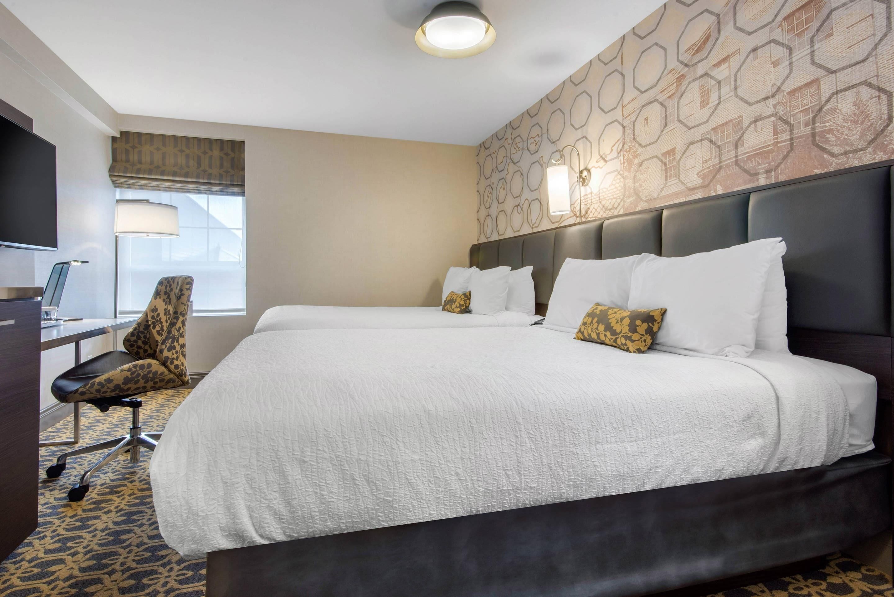 A hotel room with a large bed, a desk with a chair, a television, and a patterned carpet.