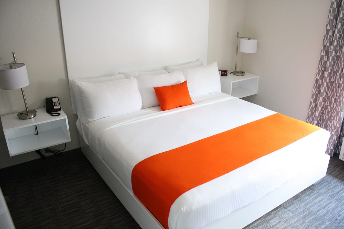 A hotel room with a large bed, white bedding, an orange throw blanket, a bedside table with a lamp, and a phone.