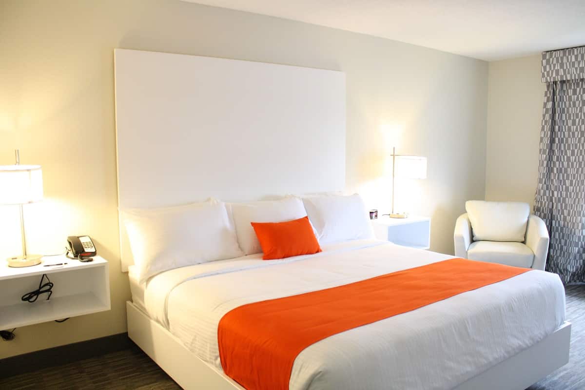 A hotel room with a large bed, white bedding, an orange throw blanket, a bedside table with a lamp, and a chair.